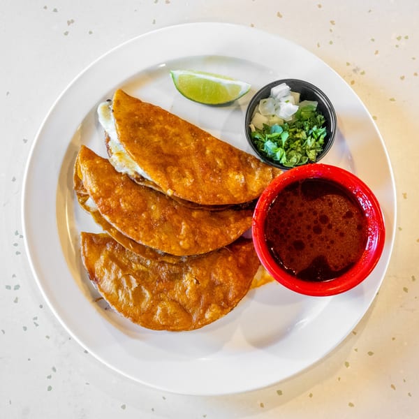 Three golden-brown birria tacos with melted cheese, a lime wedge, cilantro, onions, and a red bowl of consommé.