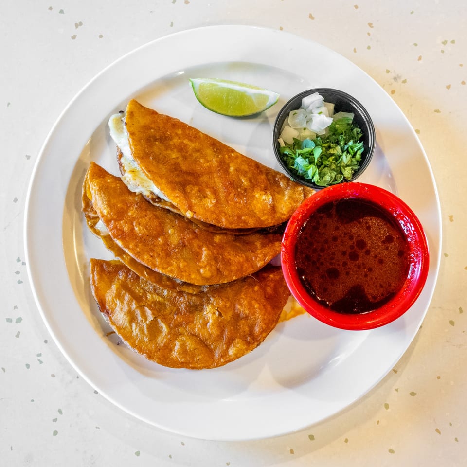 Three golden-brown birria tacos with melted cheese, a lime wedge, cilantro, onions, and a red bowl of consommé.