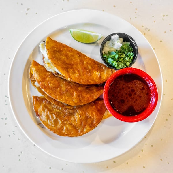 Three golden-brown birria tacos with melted cheese, a lime wedge, cilantro, onions, and a side of consommé.
