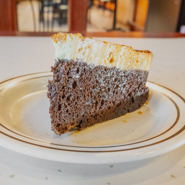 A slice of chocolate cake with a creamy white topping, dusted with powdered sugar, on a white plate.