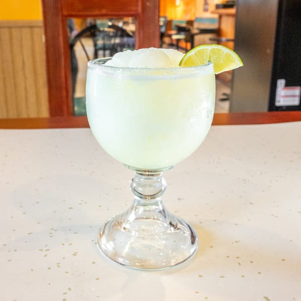 A frosty margarita with a salted rim and a lime wedge sits on a speckled white table.
