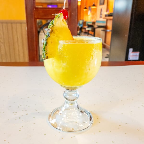 A vibrant yellow frozen drink in a stemmed glass, garnished with a pineapple slice and cherry.