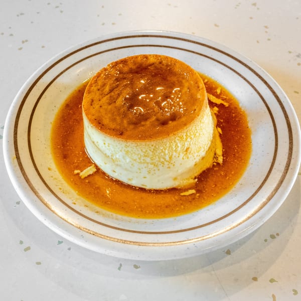 A creamy flan dessert with glistening caramel sauce on a white plate with gold stripes.