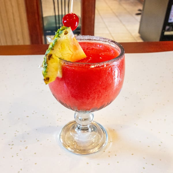 A vibrant red frozen drink in a stemmed glass, garnished with a pineapple slice and a cherry.