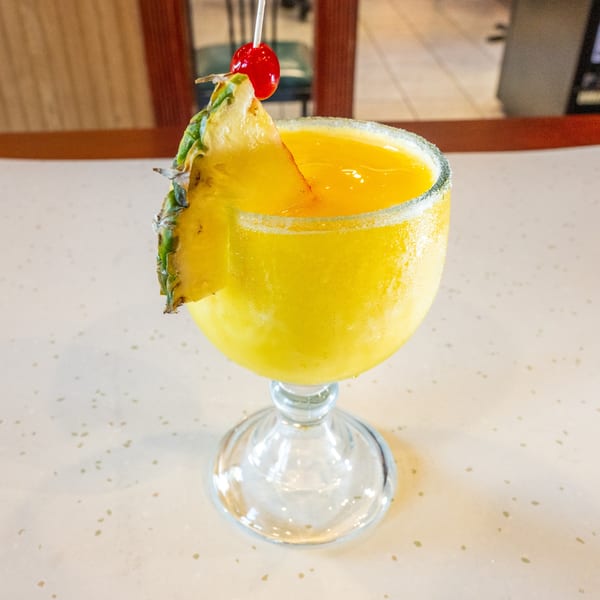 A vibrant yellow frozen drink in a stemmed glass, garnished with a pineapple slice and a cherry.