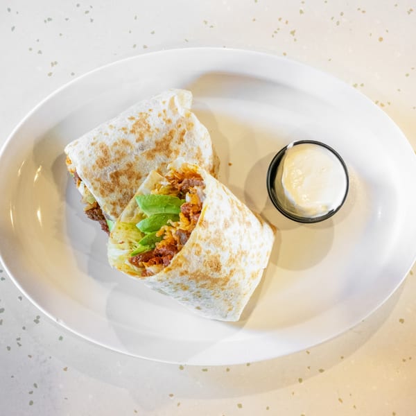 Two burritos filled with meat, lettuce, and avocado, served with a side of white sauce.