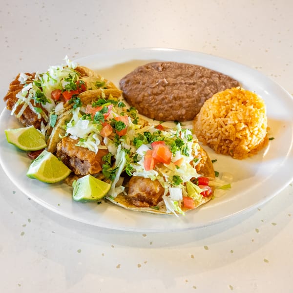 Three fish tacos with shredded lettuce, diced tomatoes, and lime wedges, served with refried beans and rice.